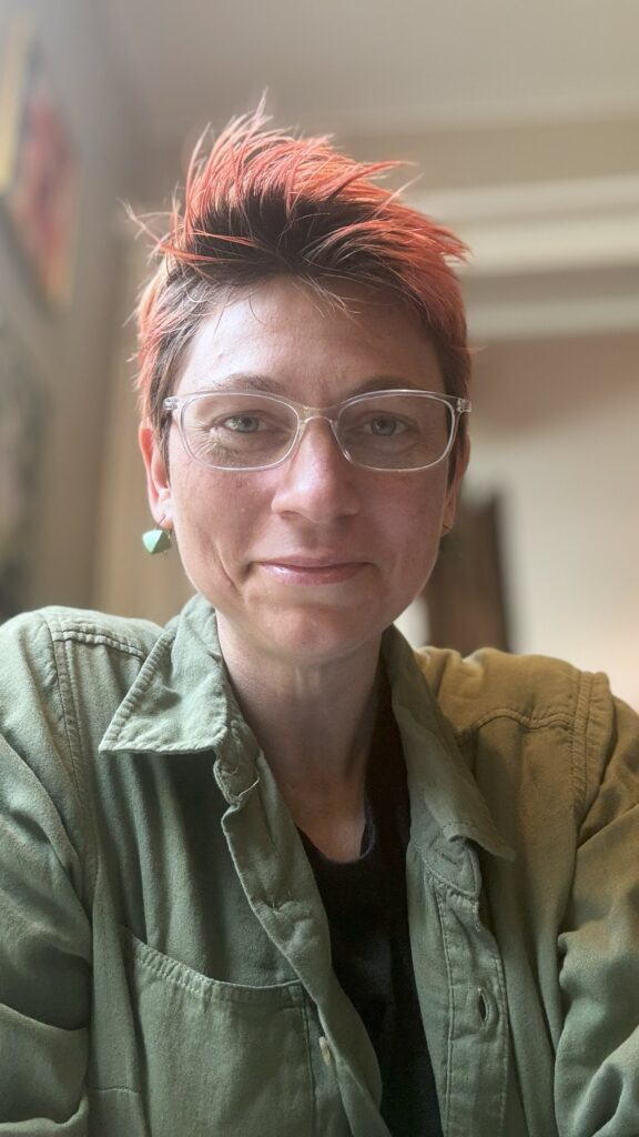 About the Artist - A Photo of Gwynneth VanLaven, seated and looking directly in the camera with wide eyes and a half-smile.  She wears clear-rimmed glasses, a green shirt with green earrings, and red hair tops her pinkish-grey complexition.